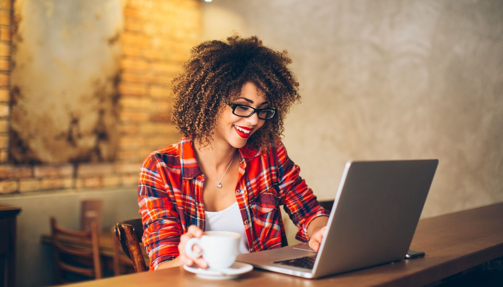 A woman with glasses and curly hair happily viewing her laptop with a cup of coffee